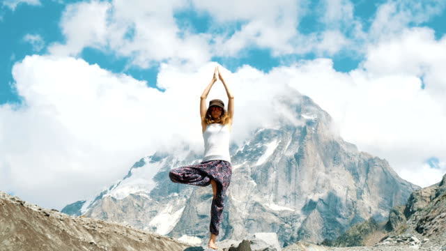 Frau-führt-einen-Asana-Baum---Vriksasana-im-Yoga-auf-einem-Hintergrund-von-einem-schneebedeckten-Berg-in-der-Wanderung.-Mädchen-macht-Gymnastik-an-der-frischen-Luft-bei-einer-Wanderung-auf-die-Natur