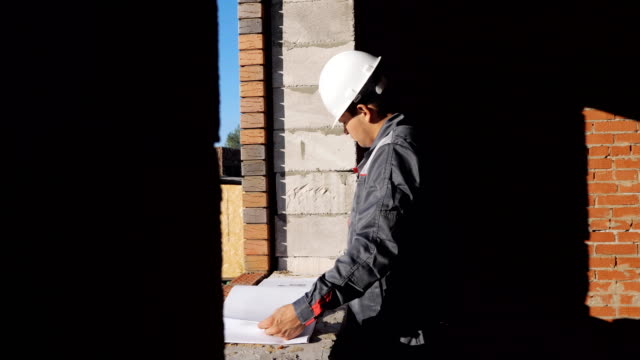Seitenansicht-des-Mannes-in-Bauarbeiterhelm-Stand-am-Fenster-im-Haus-im-Bau-Blick-auf-Papier-Entwurf,-4k