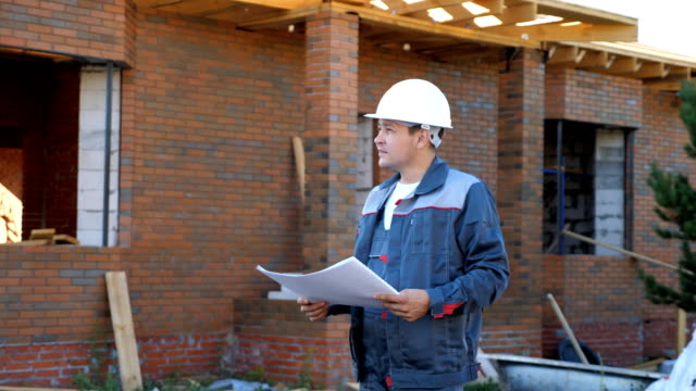 Adult-man-working-on-site-of-modern-house-and-standing-outdoors-with-paper-draft-looking-away