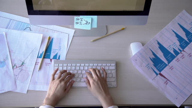 Young-female-employee-is-working,-uses-computer,-sits-at-table,-types-text,-looks-at-monitor,-presses-on-keyboard-with-fingers,-documents-with-graphs-and-diagrams-on-desk-FullHD