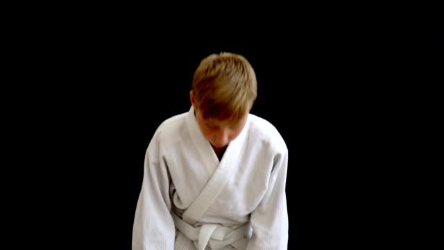 A-young-athlete-in-a-white-kimono-stands-on-a-dark-background.-The-boy-has-blond-hair-and-a-European-look.-Hands-fall,-around-the-waist-there-is-a-white-belt