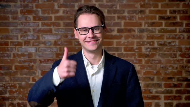 Happy-caucasian-businessman-in-glasses-demonstrating-like-gesture-and-looking-at-camera-straight,-brick-wall-behind
