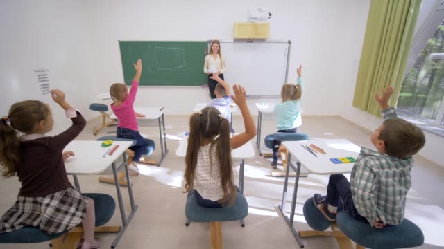Gruppe-von-Schulkindern-erhöhen-Hände-auf-Lektion-Antworten-beim-Sitzen-am-Schreibtisch-vor-Lehrer-an-Bord-in-der-Grundschule