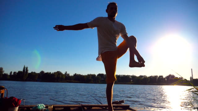 Young-guy-practicing-yoga-position-on-the-edge-of-wooden-jetty-at-lake-on-summer-day.-Sporty-man-doing-exercise-near-lake-with-sunlight-at-background.-Healthy-active-lifestyle.-Slow-motion-Close-up