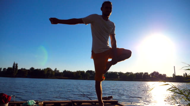 Young-guy-practicing-yoga-position-on-the-edge-of-wooden-jetty-at-lake-on-summer-day.-Sporty-man-doing-exercise-near-lake-with-sunlight-at-background.-Healthy-active-lifestyle.-Slow-motion-Close-up