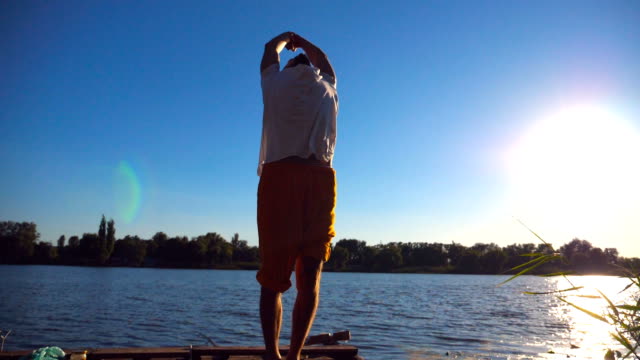 Sporty-man-standing-at-yoga-pose-on-the-edge-of-wooden-jetty-at-lake.-Young-athlete-doing-stretch-exercise-at-nature-on-sunny-summer-day.-Concept-of-healthy-active-lifestyle.-Slow-motion-Close-up