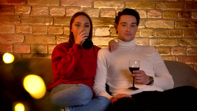 Portrait-of-young-caucasian-couple-sitting-on-sofa-and-drinking-wine-attentively-watching-movie-in-christmas-atmosphere.