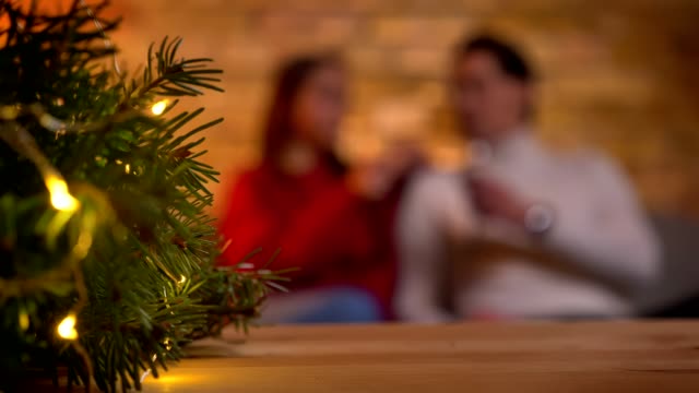 Weihnachtsbaum-mit-hellen-Lichtern-und-unscharfen-junges-Paar-auf-Sofa-sitzen-und-reden-auf-Hintergrund.