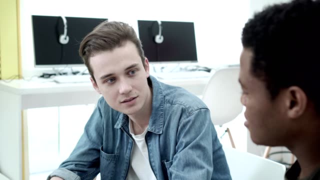 Handsome-Caucasian-student-talking-to-his-African-American-friend-while-sitting-at-desk-in-computer-classroom
