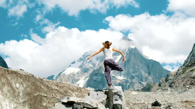 Frau-führt-einen-grundlegende-Asana-Baum---Vriksasana-im-Yoga-auf-einem-Hintergrund-von-einem-schneebedeckten-Berg-in-der-Wanderung.-Mädchen-macht-Gymnastik-an-der-frischen-Luft-bei-einer-Wanderung-auf-die-Natur