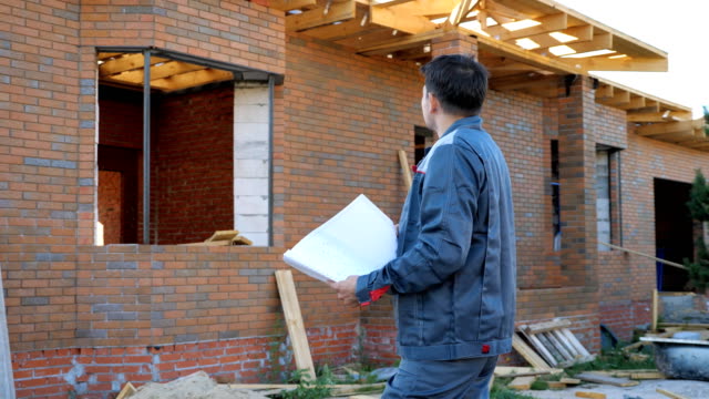 Adult-man-working-on-site-of-modern-house-and-standing-outdoors-with-paper-draft-looking-away.