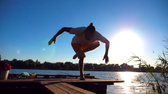 Young-guy-practicing-yoga-moves-and-positions-on-edge-of-wooden-jetty-at-lake-on-sunny-day.-Sporty-man-training-at-nature-with-sunlight-at-background.-Healthy-active-lifestyle.-Slow-motion-Close-up