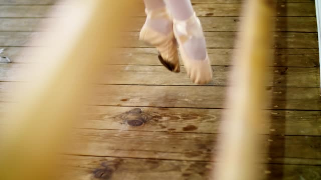 close-up,-in-dancing-hall,-ballerina-performs-pas-soute,-jumping-in-ballet-shoes-near-barre,-on-an-old-wooden-floor,-in-ballet-class