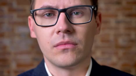 Close-up-bored-face-of-caucasian-businessman-in-black-glasses-looking-straight-and-confidently-in-brick-studio
