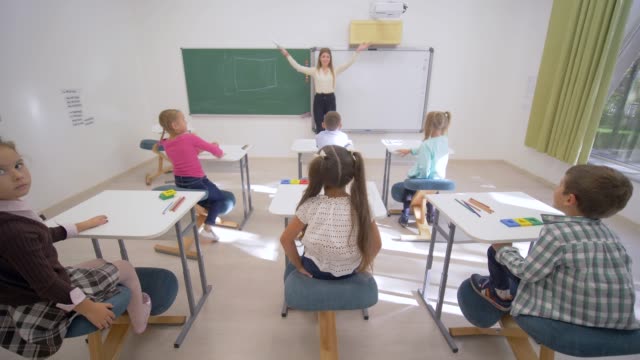 Gruppe-von-Schülern-heben-die-Hand-zur-Beantwortung-an-Lektion-beim-Sitzen-am-Schreibtisch-vor-Erzieher-Blackboard-in-Grundschule