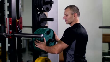 Handsome,-strong-man-puts-a-disk-on-the-barbell,-turns-to-the-camera-and-smiles-4K-Slow-Mo