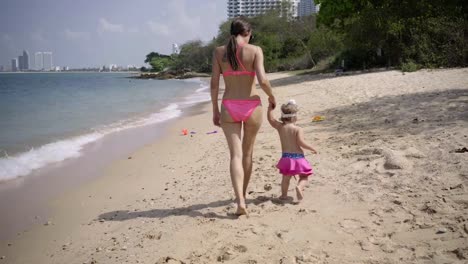 Mom-and-daughter-walk-along-the-seaside-along-the-sand-in-pink-swimsuits.-4K