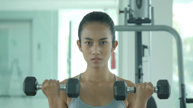 Attractive-shape-young-sporty-focused-fitness-asian-girl-with-ponytail-doing-biceps-exercises-while-sitting-on-the-bench-and-raising-dumbell-in-the-gym