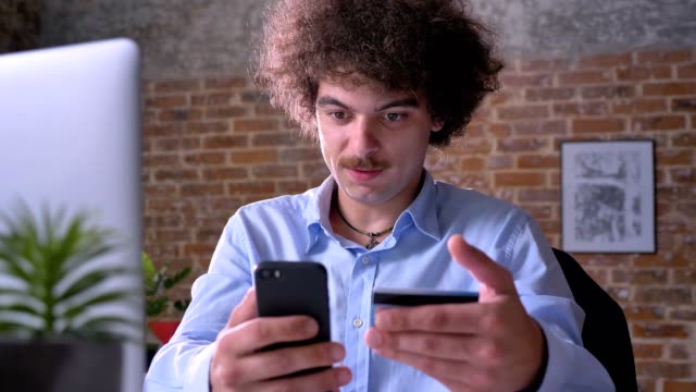 Young-concentrated-man-with-curly-hair-and-mustache-using-credit-card-and-holding-phone-sitting-at-table-in-modern-office