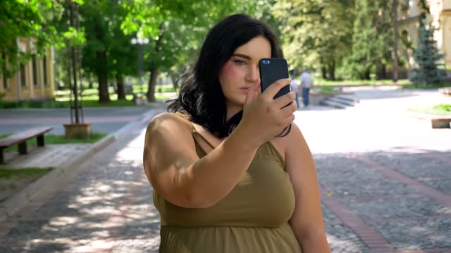 Beautiful-young-overweight-woman-taking-selfie-with-her-phone-and-doing-different-faces-in-camera,-standing-on-street-in-park