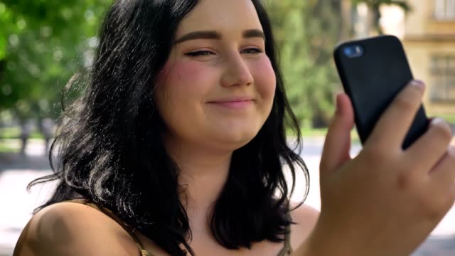 Portrait-of-young-beautiful-woman-with-obesity-taking-selfie-with-her-phone,-smiling-and-standing-on-street-in-park,-happy