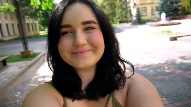 Beautiful-obese-young-woman-recording-video-on-her-web-camera-and-smiling,-standing-on-street-in-park-near-college-building