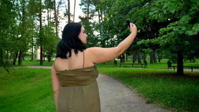 Vista-posterior-de-mujer-joven-hermosa-obesa-tomando-selfie-y-caminar-en-el-parque-con-árboles-y-hierba-verde,-encantadora-dama-de-vestido