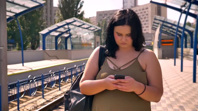 Ernste-junge-übergewichtige-Frau-mit-Rucksack-auf-Telefon-Tippen,-auf-der-Plattform-stehen-und-Zug-warten