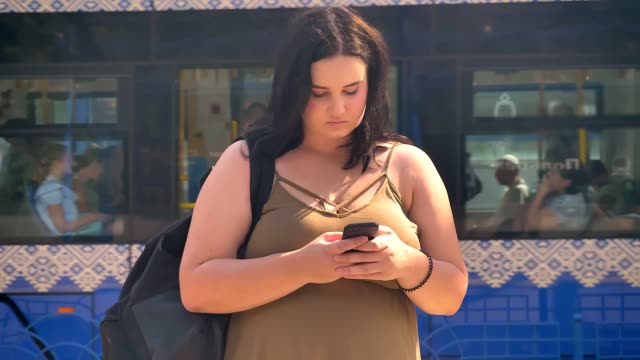 Young-serious-beautiful-obese-woman-typing-on-phone-and-standing-on-station,-train-in-background-going-away