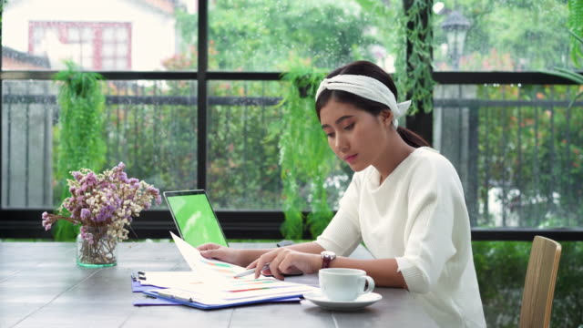 Beautiful-young-smiling-asian-woman-working-on-laptop-while-sitting-in-living-room-at-home.-Asian-business-woman-working-document-finance-and-calculator-in-her-home-office.-Enjoying-time-at-home.
