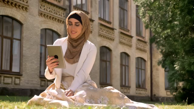 Young-muslim-woman-in-hijab-sitting-on-grass-and-having-video-chat-through-tablet,-showing-location-to-somebody,-building-in-background