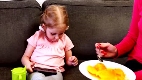 Ein-kleines-Mädchen-spielt-mit-ihrem-Smartphone,-während-sie-Obst-gefüttert-wird