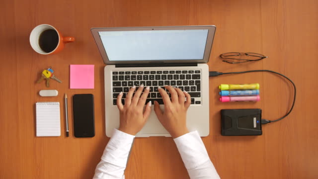 Frau-am-Schreibtisch-aus-Holz-Tippen-auf-Laptop-mit-Handy,-Kaffeetasse,-Schlüssel,-Brille,-Notebook,-externe-Festplatte-und-Marker