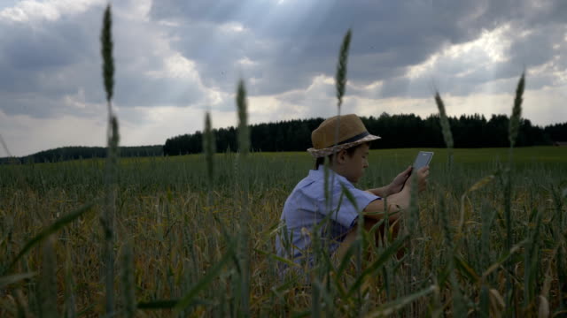 boy-sits-in-a-field-against-beautiful-clouds-and-uses-a-tablet-in-the-evening,-outdoors