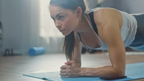 Strong-Beautiful-Fitness-Girl-in-Athletic-Workout-Clothes-is-Making-a-Plank-Exercise-While-Using-a-Stopwatch-on-Her-Phone.-She-is-Training-at-Home-in-Her-Living-Room-with-Cozy-Interior.