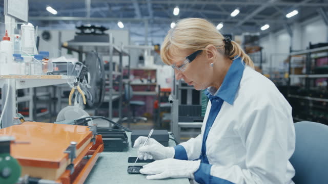 Senior-Electronics-Factory-Workers-in-White-Work-Coats-Inserting-Microchips,-Processors-and-Semiconductors-into-Printed-Circuit-Boards-for-Smartphones.-High-Tech-Factory-Facility.-Footage-with-Time-Lapse-Frames.