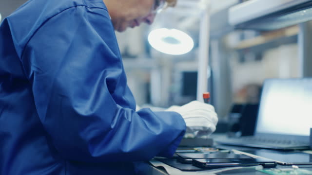 Woman-Electronics-Factory-Worker-in-Blue-Work-Coat-and-Protective-Glasses-is-Assembling-Smartphones-with-Screwdriver.-High-Tech-Factory-Facility-with-more-Employees-in-the-Background.