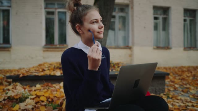 Close-up-pretty-hipster-teen-girl-sitting-on-a-sidewalk-on-autumn-city-street-and-working-laptop-computer.-Schoolgirl-using-notebook-outdoor.-Beautiful-autumn-weather