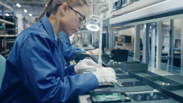 Female-Electronics-Factory-Worker-in-Blue-Work-Coat-and-Protective-Glasses-is-Assembling-Smartphones-with-Screwdriver.-High-Tech-Factory-Facility-with-more-Employees-in-the-Background.