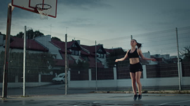 Beautiful-Energetic-Fitness-Girl-Skipping/Jumping-Rope.-She-is-Doing-a-Workout-in-a-Fenced-Outdoor-Basketball-Court.-Evening-Footage-After-Rain-in-a-Residential-Neighborhood-Area.