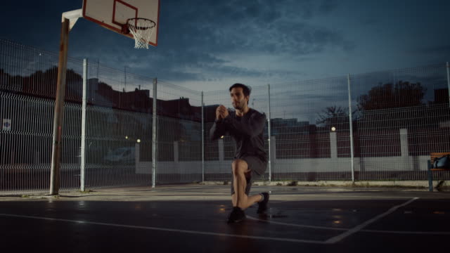 Strong-Muscular-Fit-Young-Man-in-Sport-Outfit-Doing-Forward-Lunge-Exercises.-He-is-Doing-a-Workout-in-a-Fenced-Outdoor-Basketball-Court.-Evening-Footage-After-Rain-in-a-Residential-Neighborhood-Area.