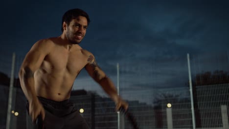 Strong-Muscular-Fit-Shirtless-Young-Man-is-Doing-Exercises-with-Battle-Ropes.-He-is-Doing-a-Workout-in-a-Fenced-Outdoor-Basketball-Court.-Evening-Footage-After-Rain-in-a-Residential-Neighborhood-Area.