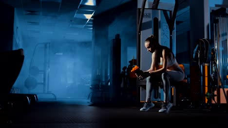 Girl-is-tired-after-training.-Sits-on-a-bench-and-drinks-from-a-bottle.-Smoke,-dark,-gym