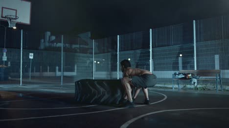Strong-Muscular-Fit-Young-Shirtless-Man-is-Doing-Exercises-in-a-Fenced-Outdoor-Basketball-Court.-He's-Flipping-a-Big-Heavy-Tire-in-a-Foggy-Night-After-Rain-in-a-Residential-Neighborhood-Area.