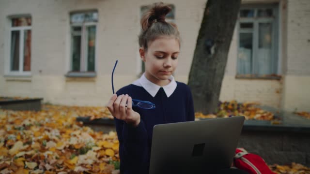 Close-up-pretty-hipster-teen-girl-sitting-on-a-sidewalk-on-autumn-city-street-and-working-laptop-computer.-Schoolgirl-using-notebook-outdoor.-Beautiful-autumn-weather