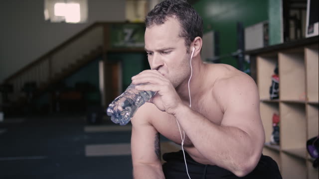 A-fit-young-man-sitting-in-a-gym-takes-a-drink-out-of-a-water-bottle-and-then-smiles-at-the-camera
