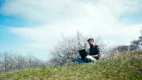 Dolly-shot.-Man-with-a-beard-uses-laptop-on-nature-near-blossoming-tree