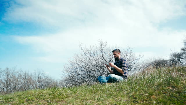 Man-with-a-beard-uses-a-tablet-on-nature-near-blossoming-tree