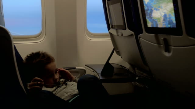 Vista-del-niño-viendo-películas-en-auriculares-en-el-avión-acostado-en-el-asiento-contra-la-ventana-del-avión