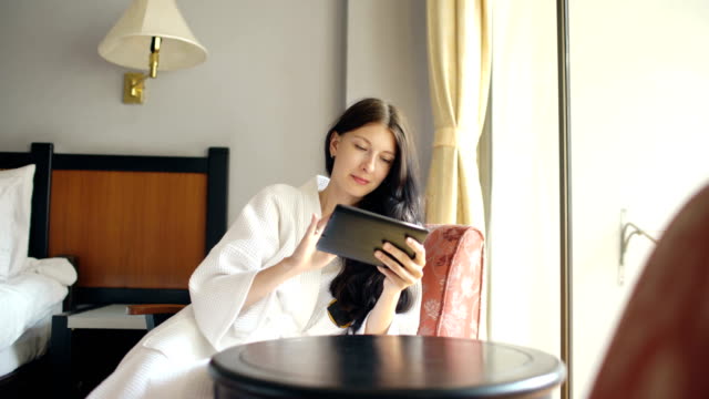 Junge-Geschäftsfrau-im-Bademantel-mit-Tablet-Computer-auf-Stuhl-im-Hotelzimmer-sitzen